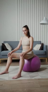 Spor kıyafetleri giymiş Avrupalı bir kız jimnastik topunda oturan kameraya bakıyor.