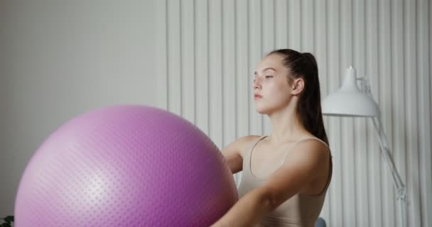 Chica europea vestida con ropa deportiva haciendo sentadillas con una pelota de gimnasio en sus manos — Vídeo de stock