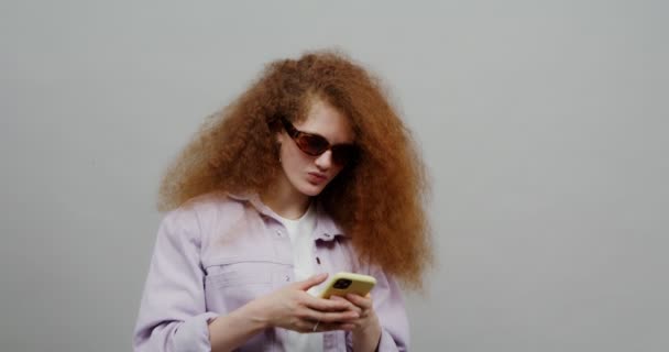 Una chica pelirroja con el pelo exuberante en gafas de sol utiliza el teléfono, mensajes de texto y riendo — Vídeo de stock