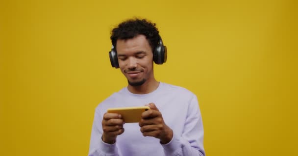 Un joven mulato con auriculares juega en el móvil y sonríe — Vídeo de stock