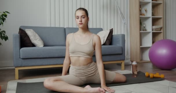 Europejska dziewczyna w sportowej odzieży patrzy w obiektyw i uśmiecha się siedząc w pół-rozłamach — Wideo stockowe
