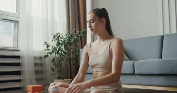 Europeisk jente i sportsvorne drikker vann fra flaske, sittende i lotusstilling – stockvideo