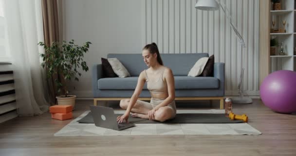 Europees meisje in sportkleding maakt gebruik van een laptop zitten in lotus positie — Stockvideo