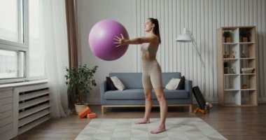 Spor kıyafetleri giymiş Avrupalı bir kız elinde spor topuyla çömelme hareketleri yapıyor.