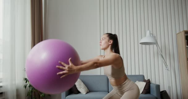 Spor kıyafetleri giymiş Avrupalı bir kız elinde spor topuyla çömelme hareketleri yapıyor. — Stok video