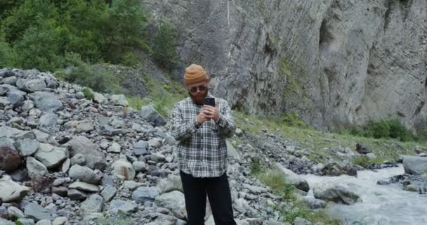 El Cáucaso. Joven toma fotos en el teléfono de pie cerca de un río de montaña — Vídeos de Stock