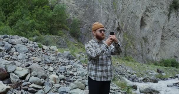 Der Kaukasus. Junger Mann fotografiert am Telefon stehend in der Nähe eines Gebirgsflusses — Stockvideo