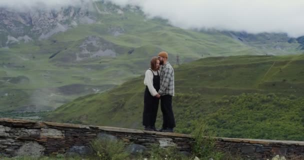 Una joven pareja se para en el parapeto abrazándose contra el telón de fondo de las montañas — Vídeos de Stock