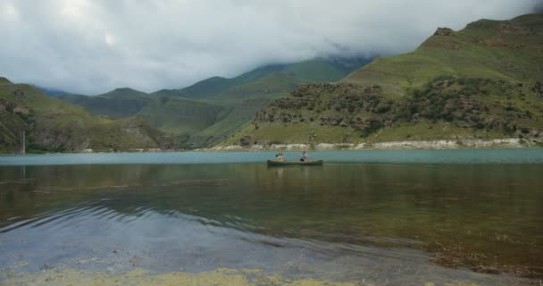 Um jovem casal está navegando em barco a remos, através do lago contra o pano de fundo das montanhas — Vídeo de Stock
