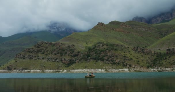 Una pareja está sentada en un bote de remos en el lago contra un telón de fondo de montañas. — Vídeo de stock