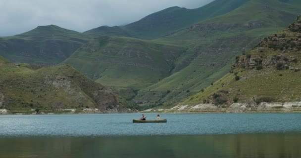 Una joven pareja está navegando en bote de remos, a través del lago contra el telón de fondo de las montañas — Vídeos de Stock