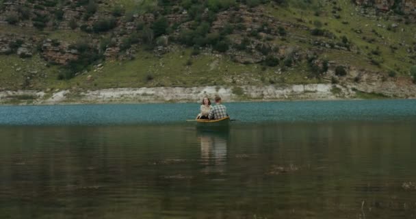 Una giovane coppia sta navigando in barca a remi, attraverso il lago sullo sfondo delle montagne — Video Stock