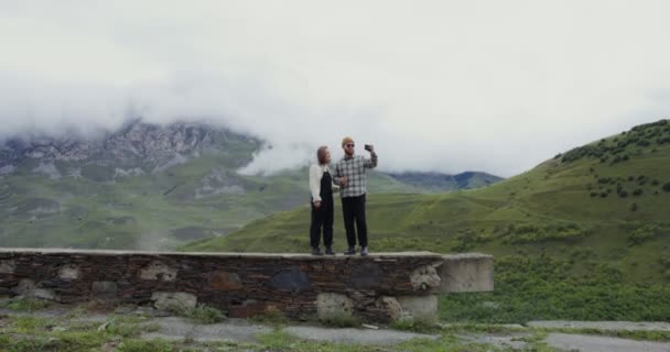 Una giovane coppia prende selfie sul cellulare, in piedi sullo sfondo delle montagne — Video Stock