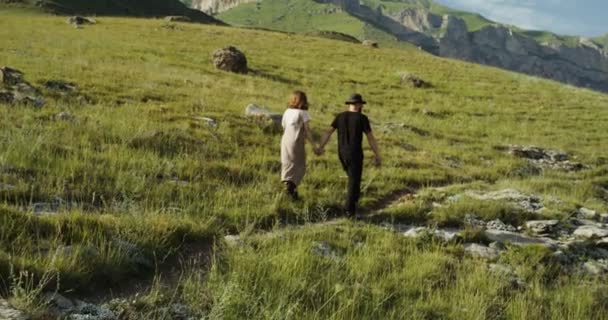 ロシア、コーカサス。若いカップルが緑の丘を登る手を握って — ストック動画