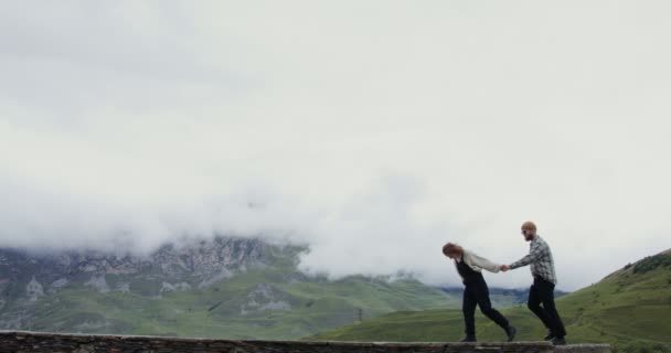 Ett ungt par gå på parapet hålla händerna mot bakgrund av berg — Stockvideo