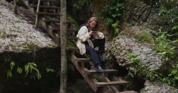 Russie, Caucase. Une femme utilise le téléphone assis sur un escalier en bois parmi les rochers — Video