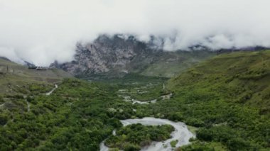 Dağlık bir vadi, ovalarda nehir ve bulutlarla kaplı kayalık dağlar.