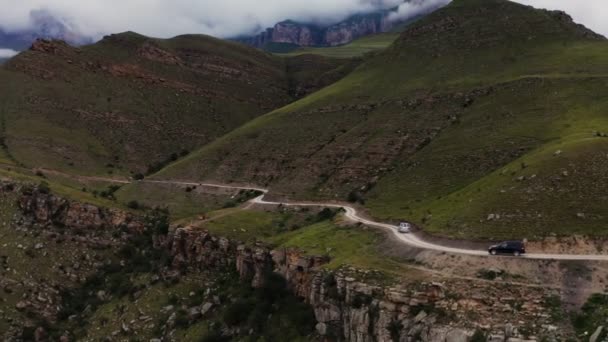 Coches conducen a lo largo de una estrecha carretera de montaña de grava por una pendiente cubierta de hierba empinada — Vídeo de stock