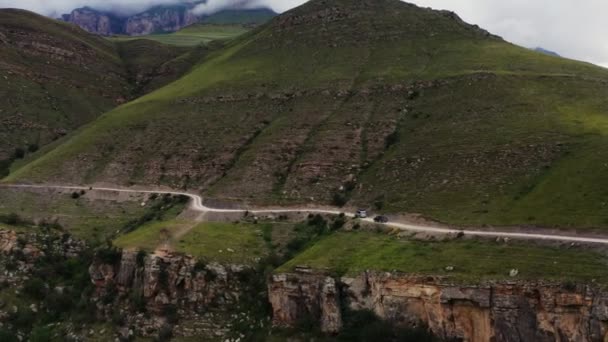 Автомобілі їдуть по вузькій гравійній гірській дорозі вгору крутим трав'янистим схилом — стокове відео
