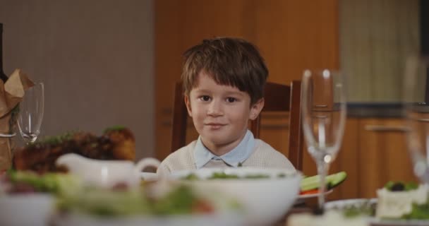 Een lachend jongetje kijkt recht naar de camera, zittend aan een gedekte feestelijke tafel — Stockvideo