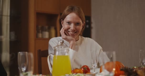Μια νεαρή γυναίκα χαμογελώντας κοιτάζει απευθείας στην κάμερα, κάθεται στο στρωμένο γιορτινό τραπέζι — Αρχείο Βίντεο