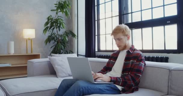 Un hombre con barba de utiliza un ordenador portátil sentado en un sofá en un interior de luz moderna — Vídeo de stock