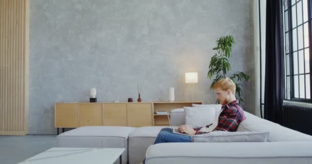 A man with a beard of uses a laptop sitting on a sofa in a modern light interior — Stock Video