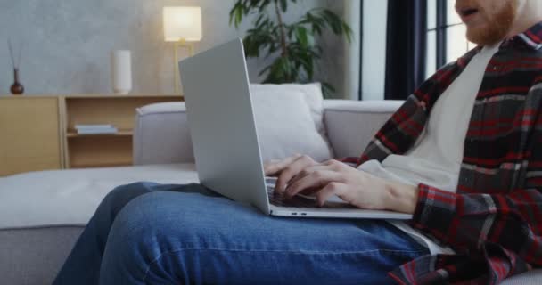 Un joven con barba termina de trabajar en una laptop, bostezando y frotándose los ojos — Vídeos de Stock