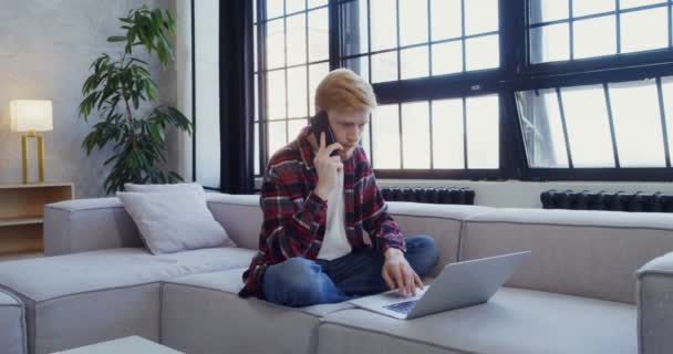 Un joven trabaja en un portátil y habla en un móvil mientras está sentado en un sofá — Vídeo de stock