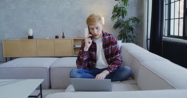 Ein junger Mann arbeitet an einem Laptop und spricht auf einem Sofa mit einem Handy — Stockvideo