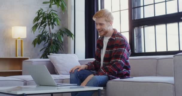 Bonito homem em fones de ouvido sorri, falando em videochamada no laptop — Vídeo de Stock