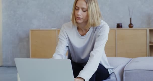 Uma jovem mulher sorrindo falando em videochamada no laptop enquanto se senta no sofá — Vídeo de Stock