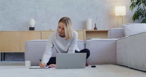 Kulaklıklı genç bir kadın dizüstü bilgisayarda çalışıyor, not defterine not alıyor. — Stok video