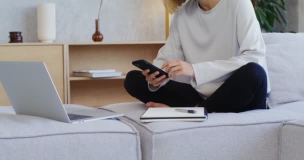 Jovem mulher sorrindo usando telefone enquanto sentado no sofá com bloco de notas e laptop — Vídeo de Stock