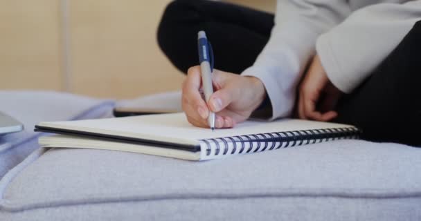 Eine Frau macht sich mit einem Stift Notizen in einem Notizbuch, sitzt vor einem offenen Laptop — Stockvideo