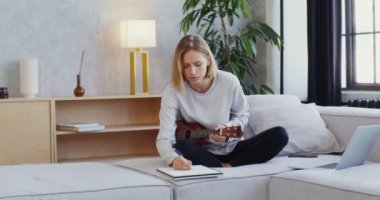 Kadın ukulele çalıyor, dizüstü bilgisayarda oturuyor ve not defterine notlar yazıyor.