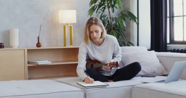 Nő játszik ukulele ül az első laptop és jegyzeteket jegyzetfüzetben — Stock videók