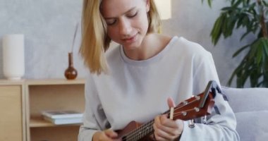 Genç bir kadın ukulele çalıyor ve kanepede oturuyor.