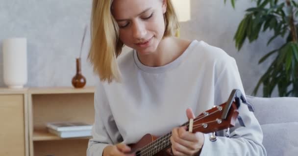 Eine junge Frau spielt Ukulele und singt, auf dem Sofa sitzend — Stockvideo