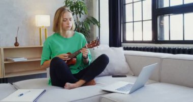 Genç bir kadın kanepede dizüstü bilgisayar ve not defterinin yanında gitar çalıyor.