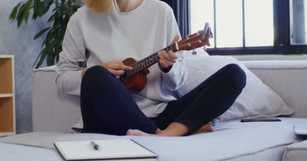 Una joven toca el ukelele sentado en el sofá junto a la computadora portátil y el bloc de notas — Vídeos de Stock