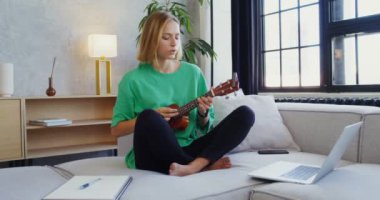 Genç bir kadın kanepede oturup bilgisayarına bakarken ukulele çalıyor.