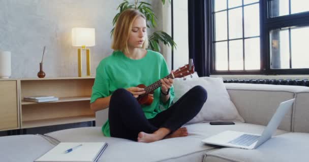 En ung kvinna spelar ukulele medan du sitter på soffan och tittar in i öppen bärbar dator — Stockvideo