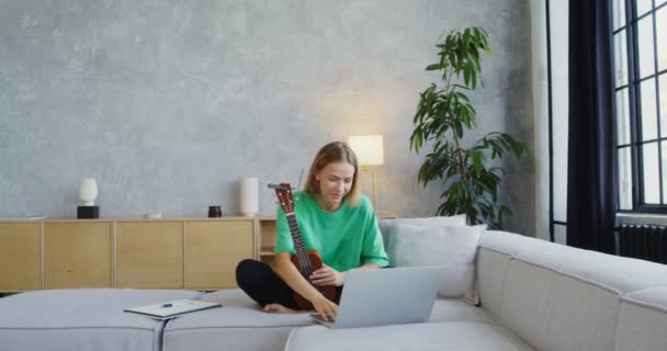 Uma jovem toca ukulele enquanto a chamada de vídeo no laptop, sentado no sofá — Vídeo de Stock