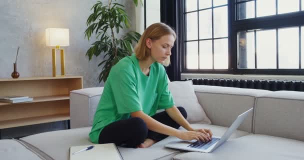 Een jonge vrouw werkt op een laptop zittend op een bank in een modern interieur — Stockvideo