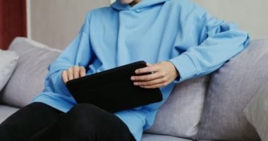 Gündelik giysiler içinde tablet kullanan, modern iç mekanda koltukta oturan genç bir kadın.