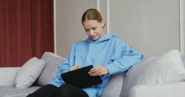 Gündelik giysiler içinde tablet kullanan, modern iç mekanda koltukta oturan genç bir kadın.