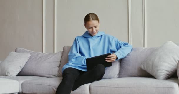 Jonge vrouw in casual kleding met behulp van tablet, zittend op de bank in een modern interieur — Stockvideo