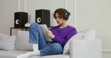 Kulaklıklı yakışıklı adam kanepede otururken not alır ya da çizer.