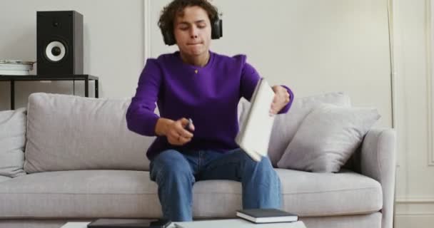 Handsome guy in headphones makes notes or draws in notebook sitting on sofa — Stock Video